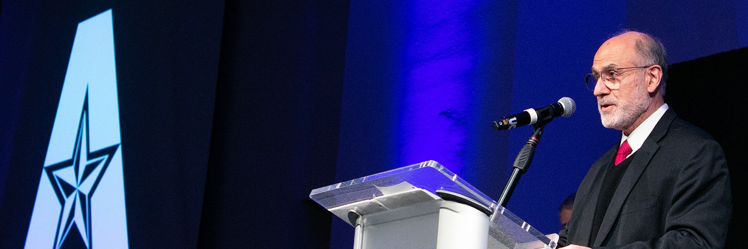 Man standing at a podium on a stage. Behind him on the wall is a large UTA logo