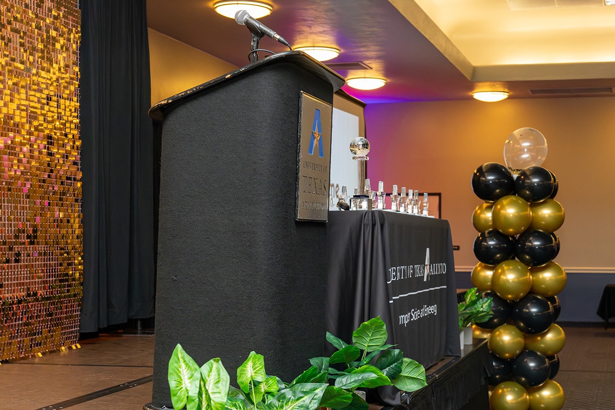 A stage with a podium, a table with trophies, green plants, black and gold balloons, and a golden sequin backdrop - opens in a new tab
